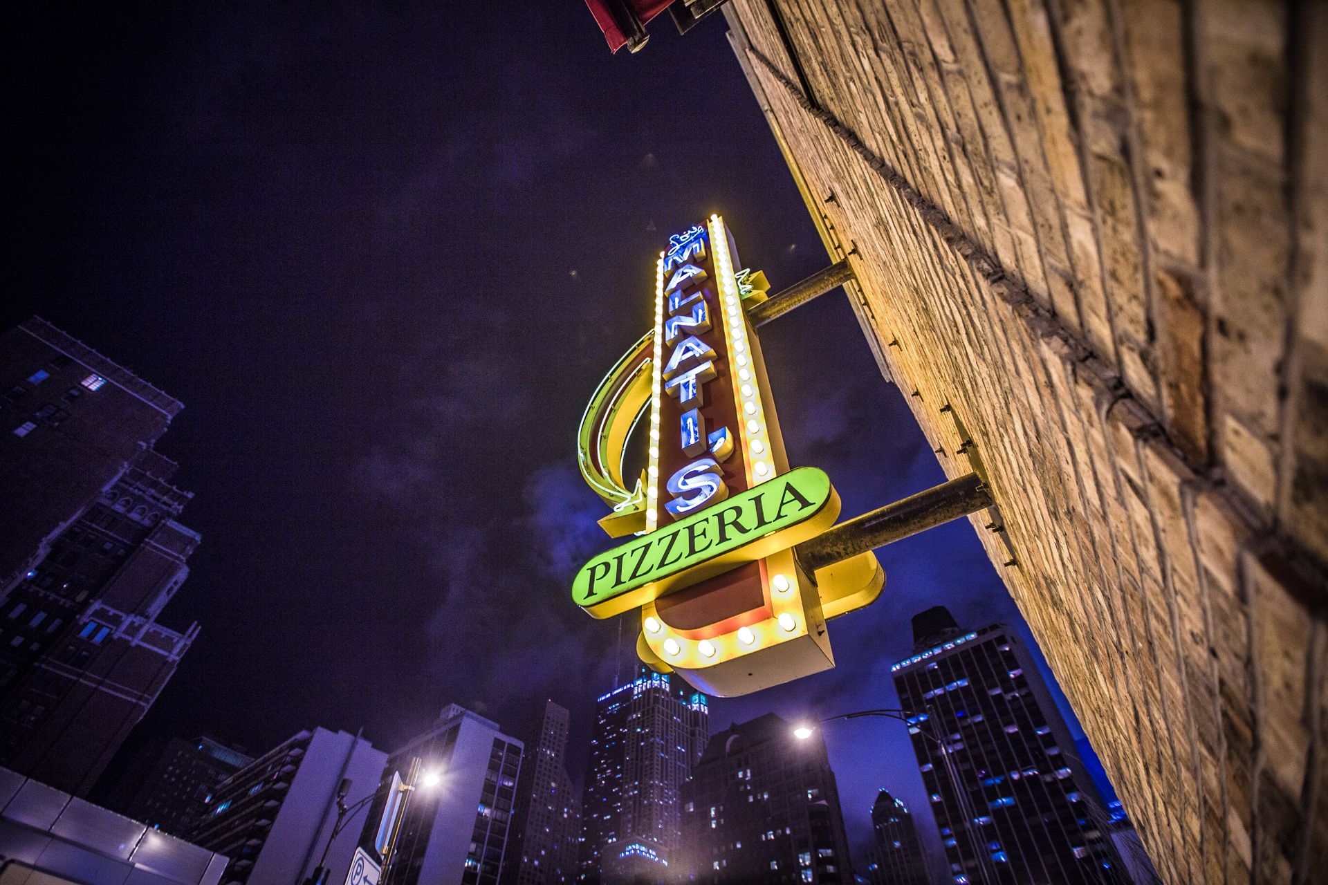 Illuminated pizzeria sign with city skyline at night, captured from a low angle.