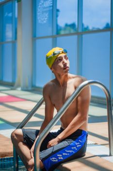 Adult male swimmer sitting by the poolside in a bright indoor venue.