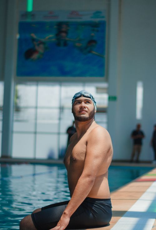 Free A male swimmer in swim gear sits with confidence at the edge of an indoor swimming pool, preparing for a swim session. Stock Photo