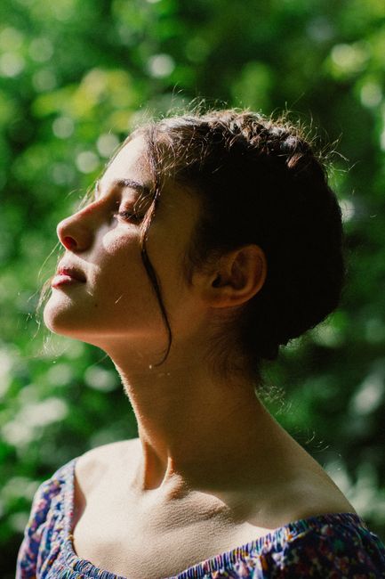 Profile of a woman with closed eyes enjoying sunlight in a lush green setting.