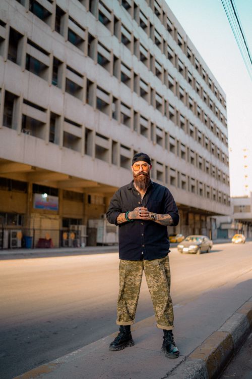 Free Bearded man stands confidently on an urban street, showcasing trendy fashion against a city facade. Stock Photo