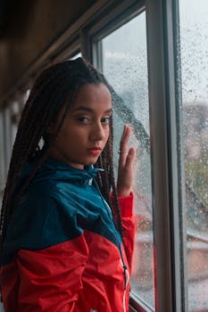 一位扎着辫子的年轻女子望着窗外的雨，肖像传达出体贴之情