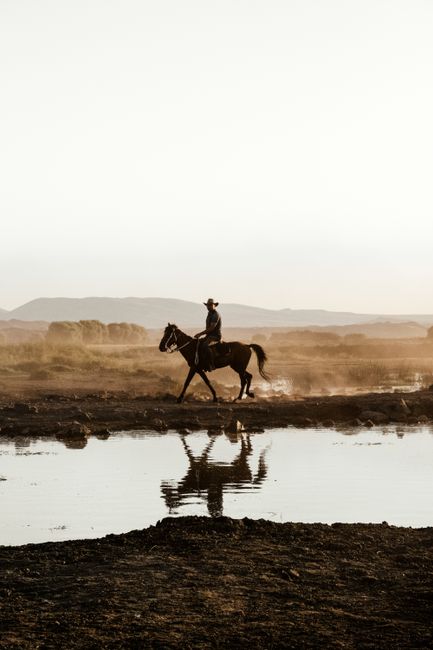 A lone rider on horseback crosses a river, reflecting on the water at sunset.
