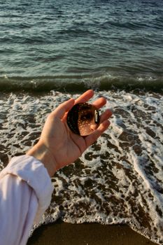 A hand holds a mirror reflecting a face at the beach with waves in the background.