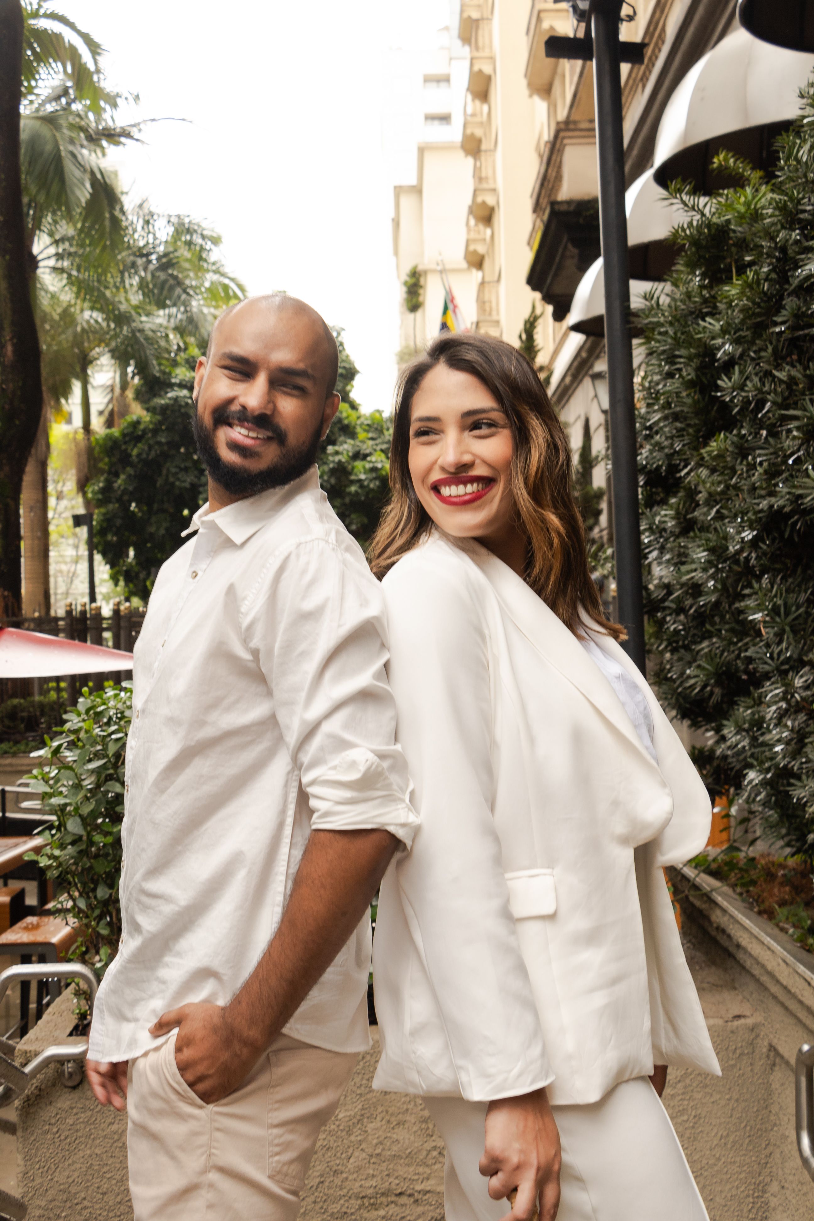 Elegant couple smiling on a São Paulo street in white outfits, showcasing urban fashion and happiness.