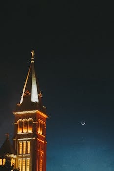 一座明亮的教堂塔楼，顶端耸立着十字架，在夜空和新月的映衬下格外醒目
