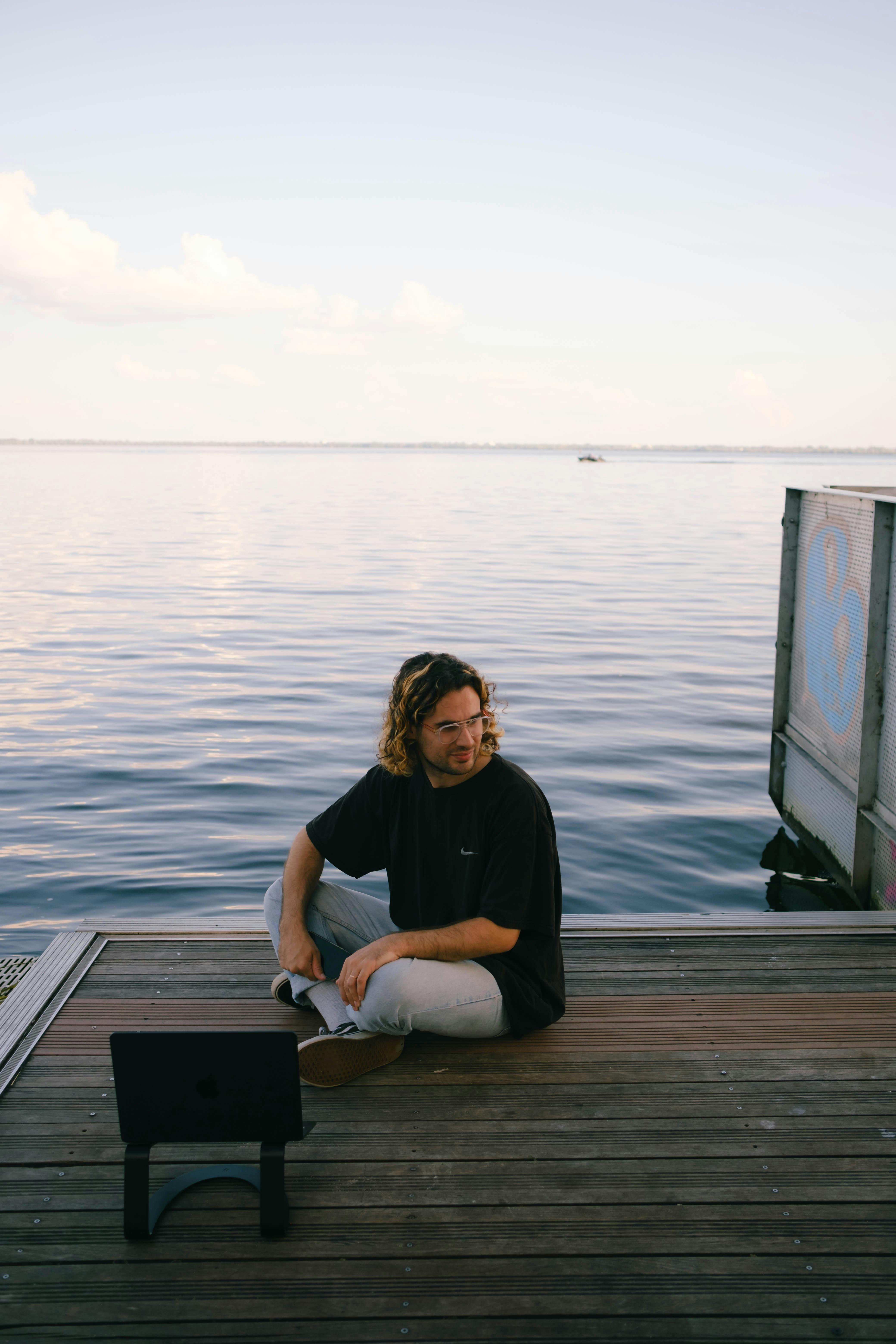 A man with long hair sits on a pier by the sea using a laptop. Tranquil outdoor scene.