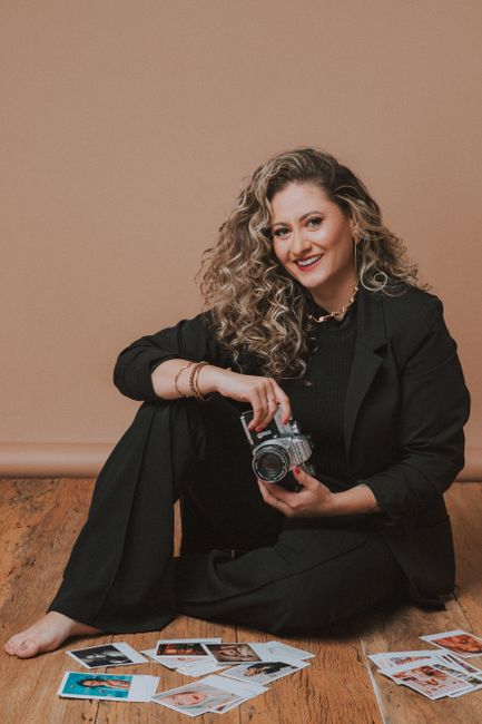 Woman in black blazer holding a camera, surrounded by polaroids, showcasing creativity and style.