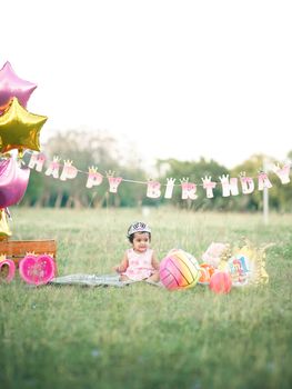 Cute baby girl enjoying her birthday in a sunny meadow with balloons and gifts.