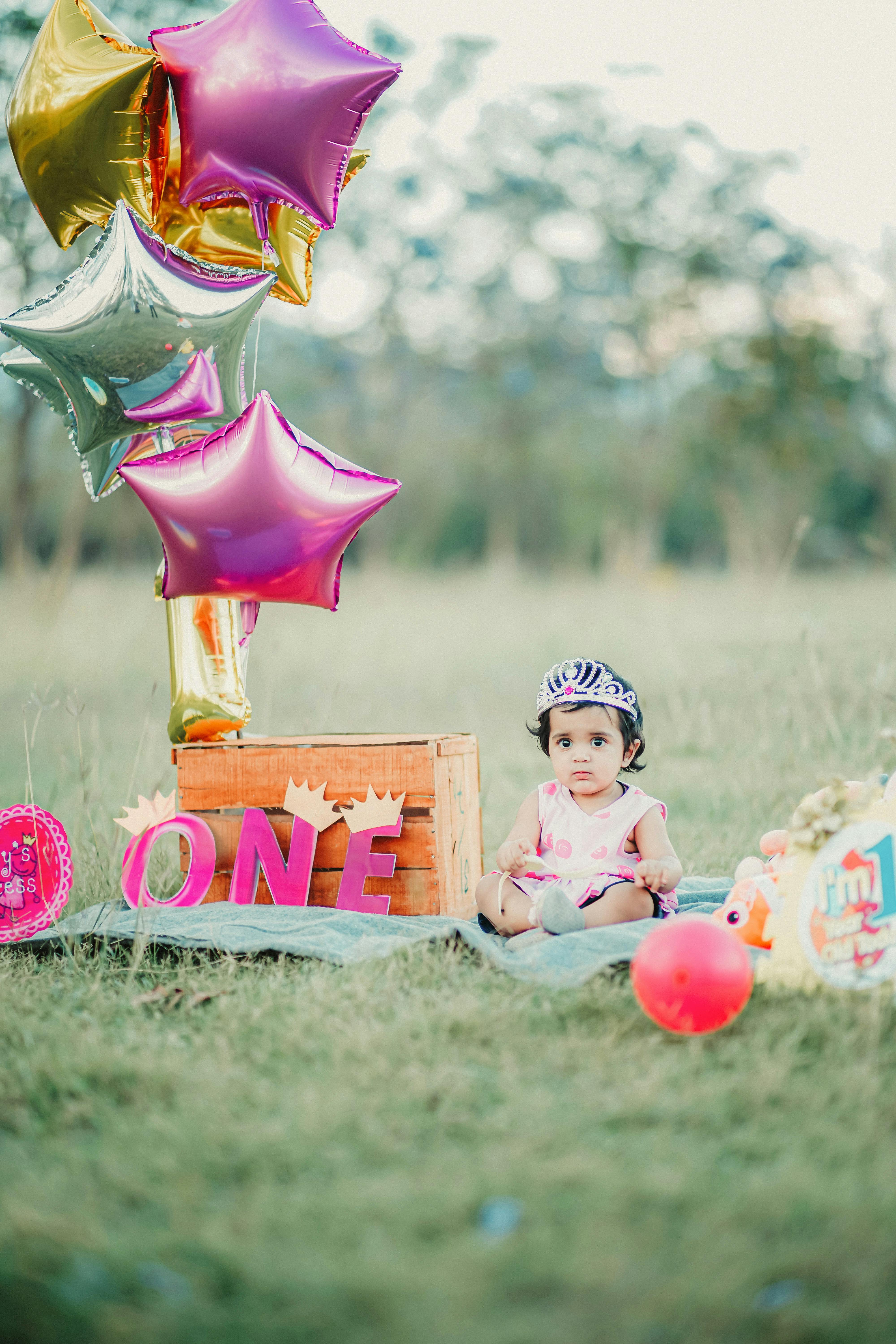 A cute baby girl with a crown sitting outdoors surrounded by balloons and birthday decorations.