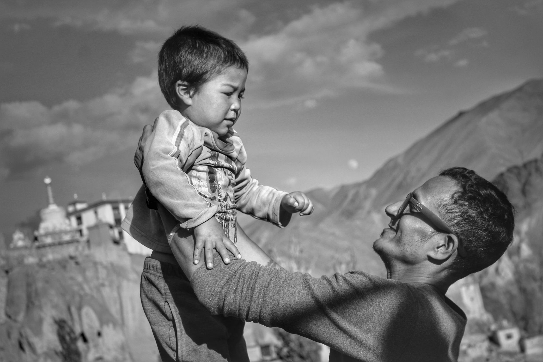 A heartwarming black and white photo of a father lifting his son in the scenic Lamayuru landscape.