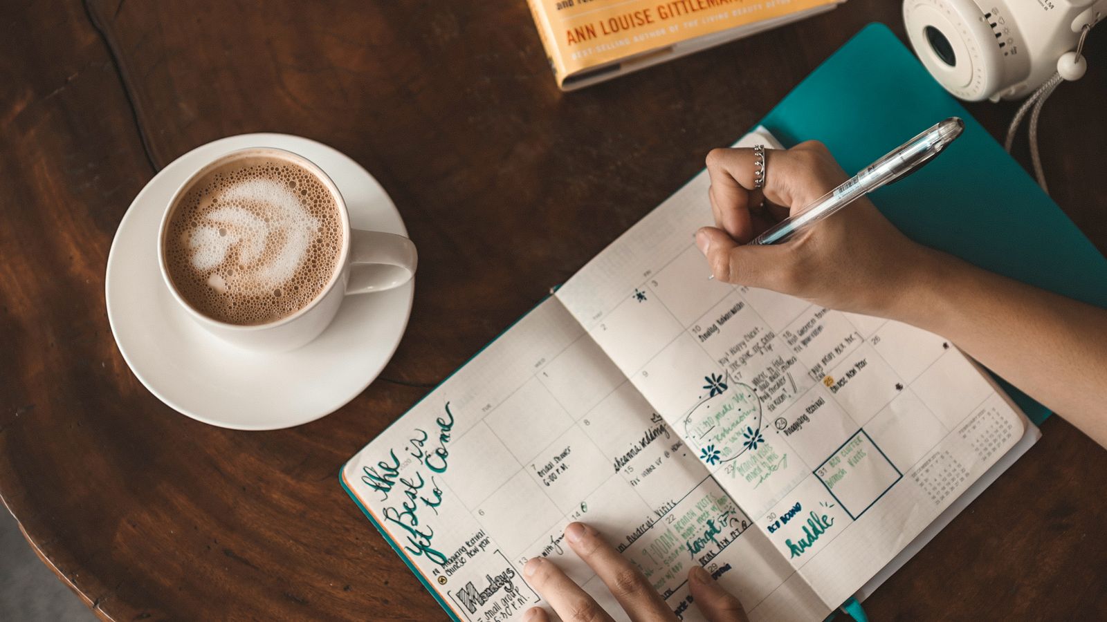 A woman writing in a planner beside a cappuccino, book and camera on a wooden table