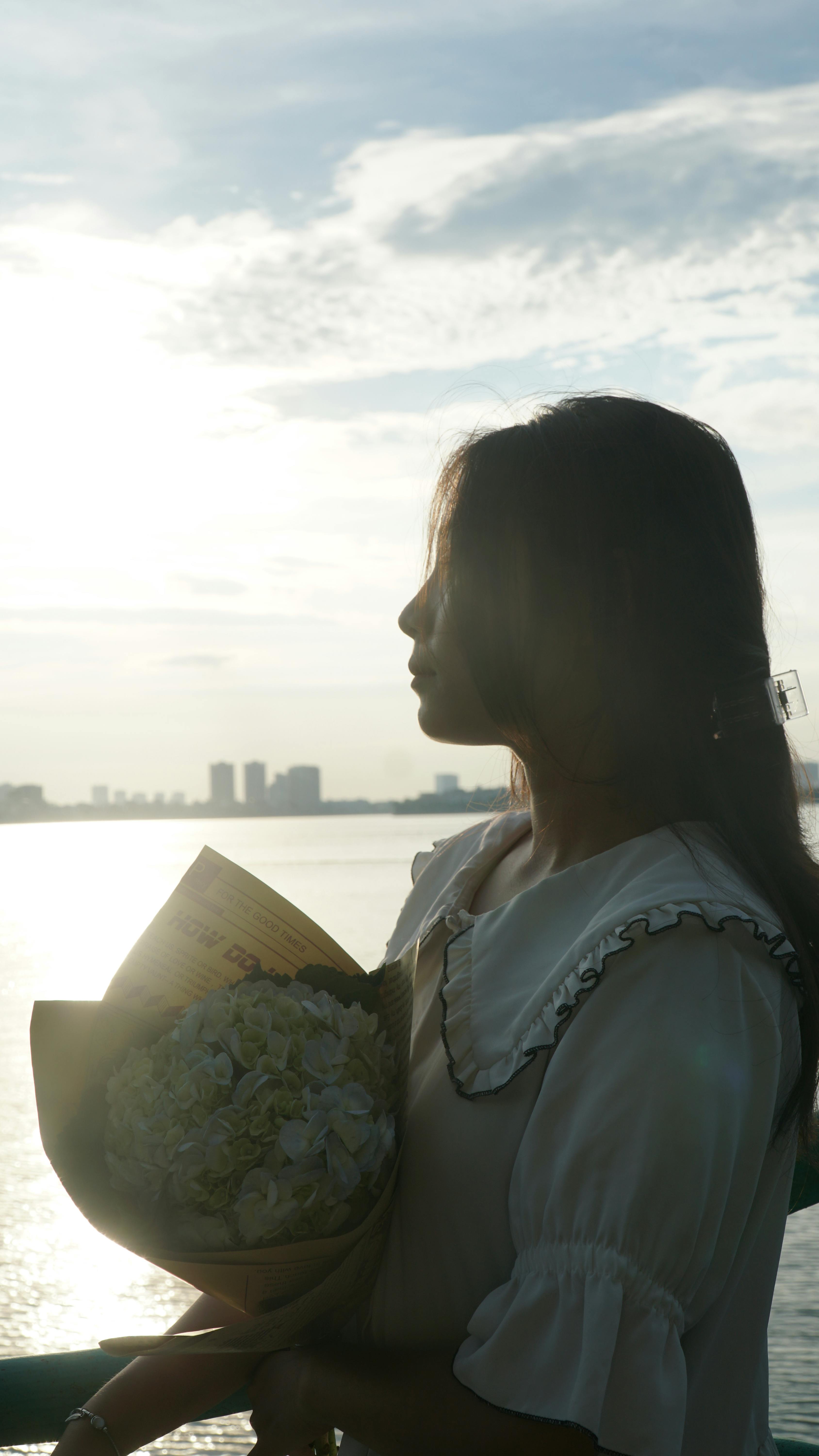 Silhouette of a woman with a bouquet, backlit by the warm glow of the sunset over a cityscape.