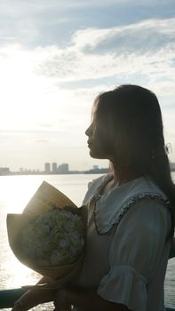 Silhouette of a woman with a bouquet, backlit by the warm glow of the sunset over a cityscape.
