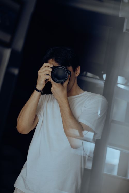 Free A man in a white shirt holds a camera, immersed in taking a photo indoors. Stock Photo