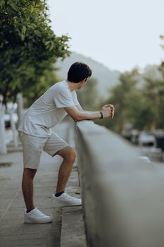 Man in white t-shirt and shorts leaning on railing outdoors with blurred background.