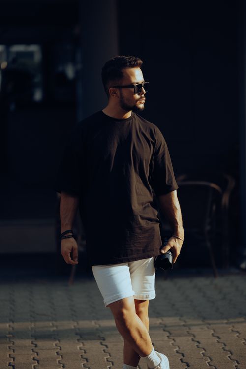 Free A fashionable man in a black t-shirt and white shorts poses confidently holding a camera outdoors. Stock Photo