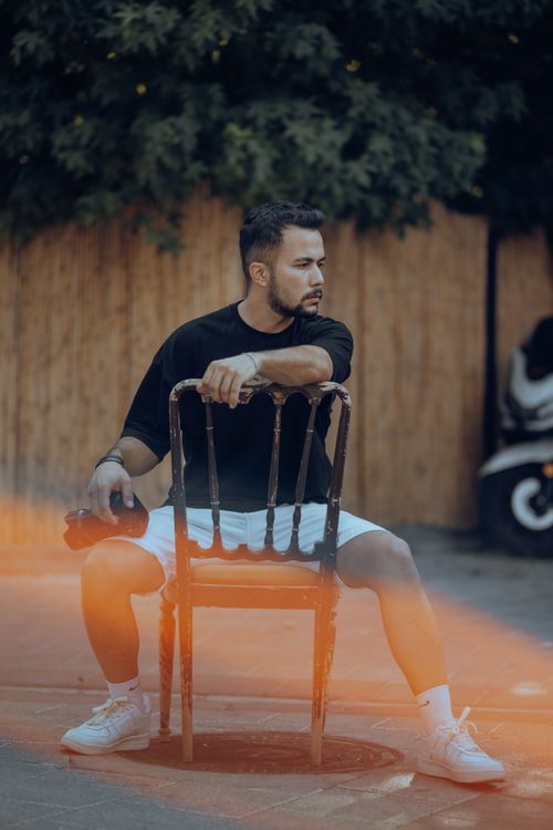Free A fashionable man posing outdoors on a wooden chair in a casual outfit, creating a cool urban vibe. Stock Photo