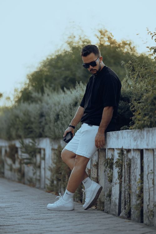 Free Fashionable man wearing sunglasses and sneakers leaning against a wall outdoors. Stock Photo