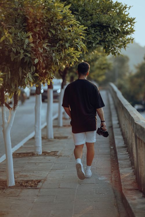 Free A man in a black t-shirt walks along a tree-lined sidewalk holding a camera, capturing the essence of street photography. Stock Photo
