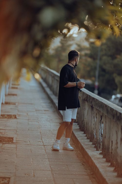 Free Man standing on bridge, enjoying the urban view during sunset, wearing a black T-shirt and shorts. Stock Photo