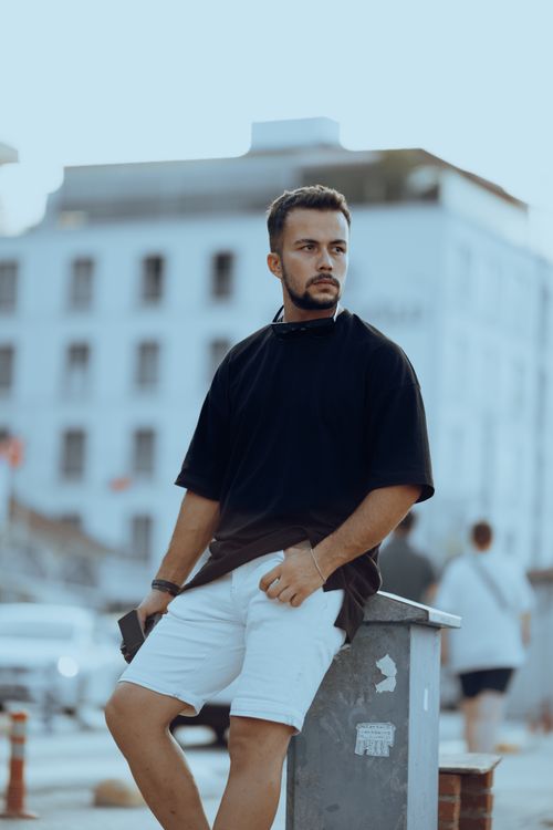 Free Fashionable man in black t-shirt and white shorts against urban backdrop, showcasing modern style. Stock Photo