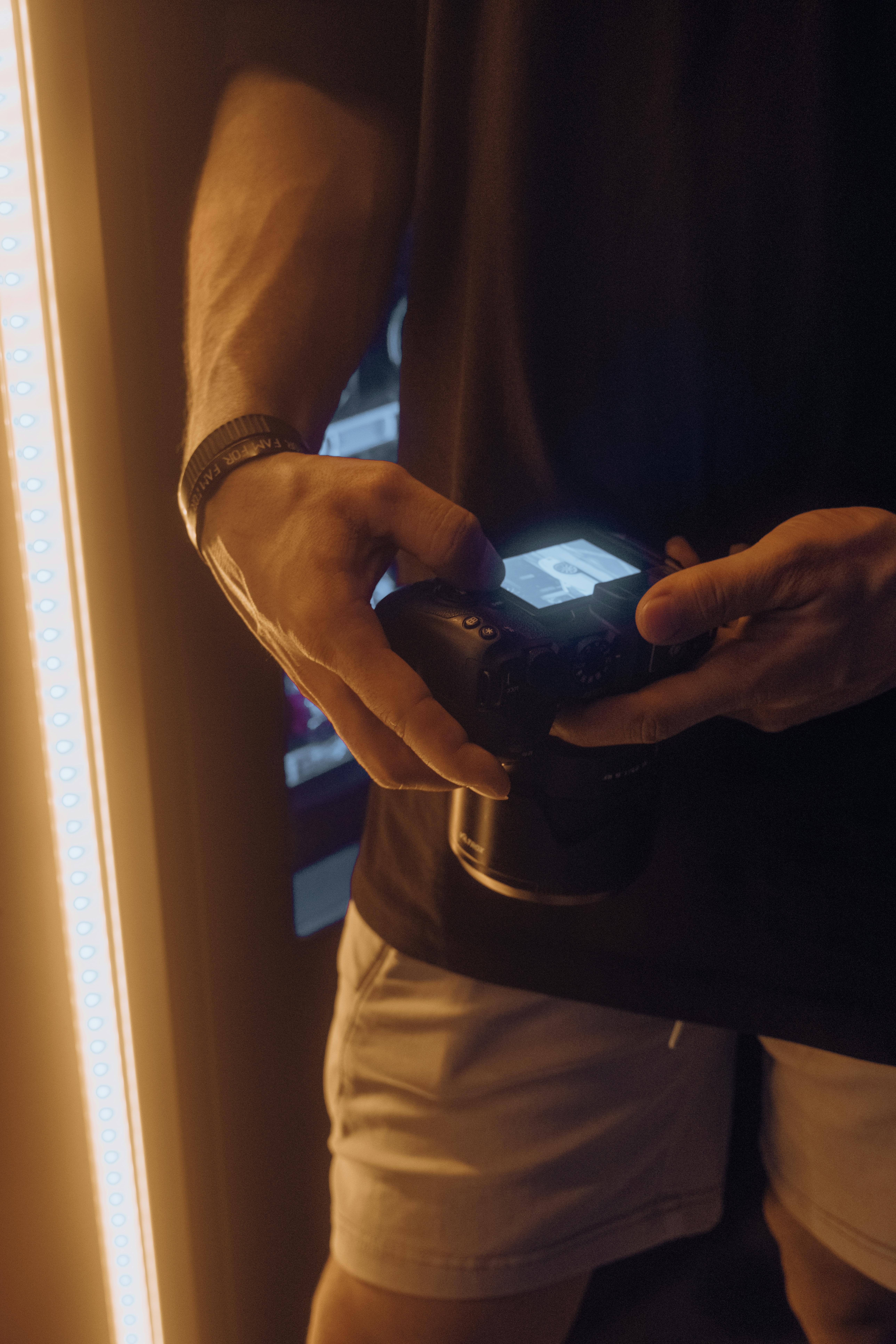 Free Close-up of a man holding a camera, adjusting settings indoors with ambient lighting. Stock Photo