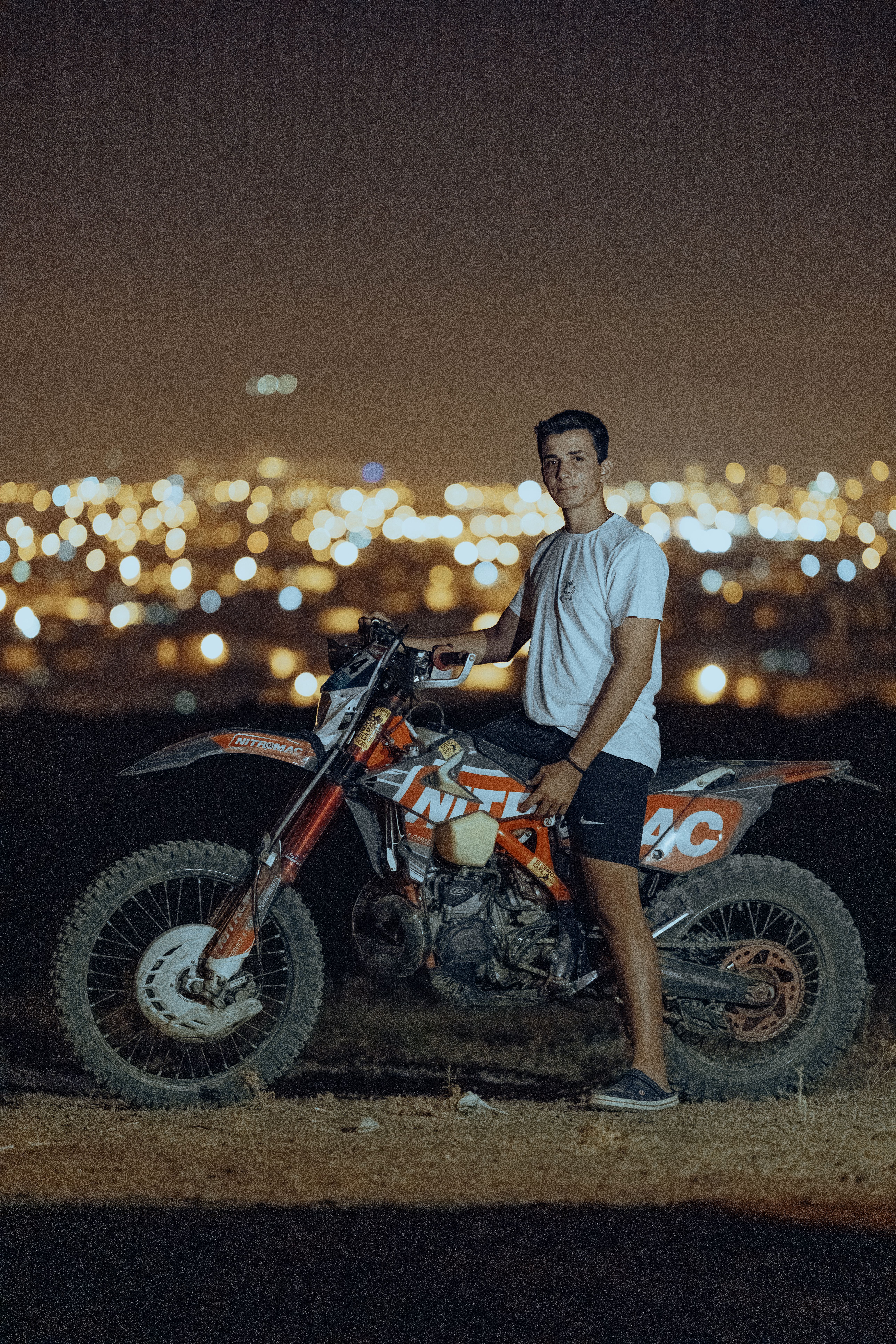A young man standing next to a motorcycle with a cityscape illuminated in the background at night.