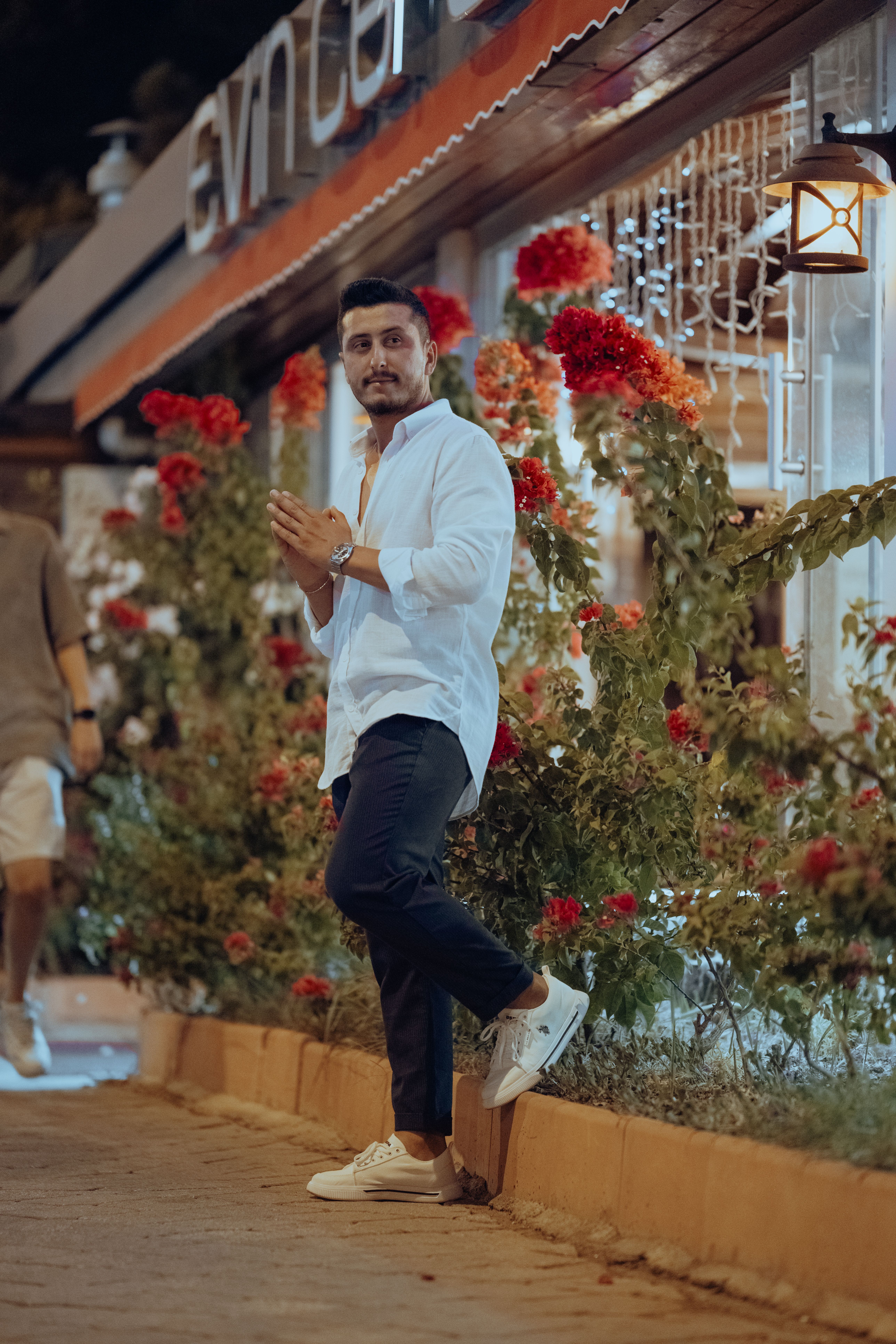A man poses casually in a white shirt by a vibrant flower garden, illuminated at night.