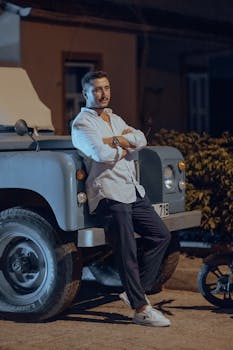 Man in white shirt leaning against a vintage car on a dimly lit street, exuding a cool and relaxed vibe.