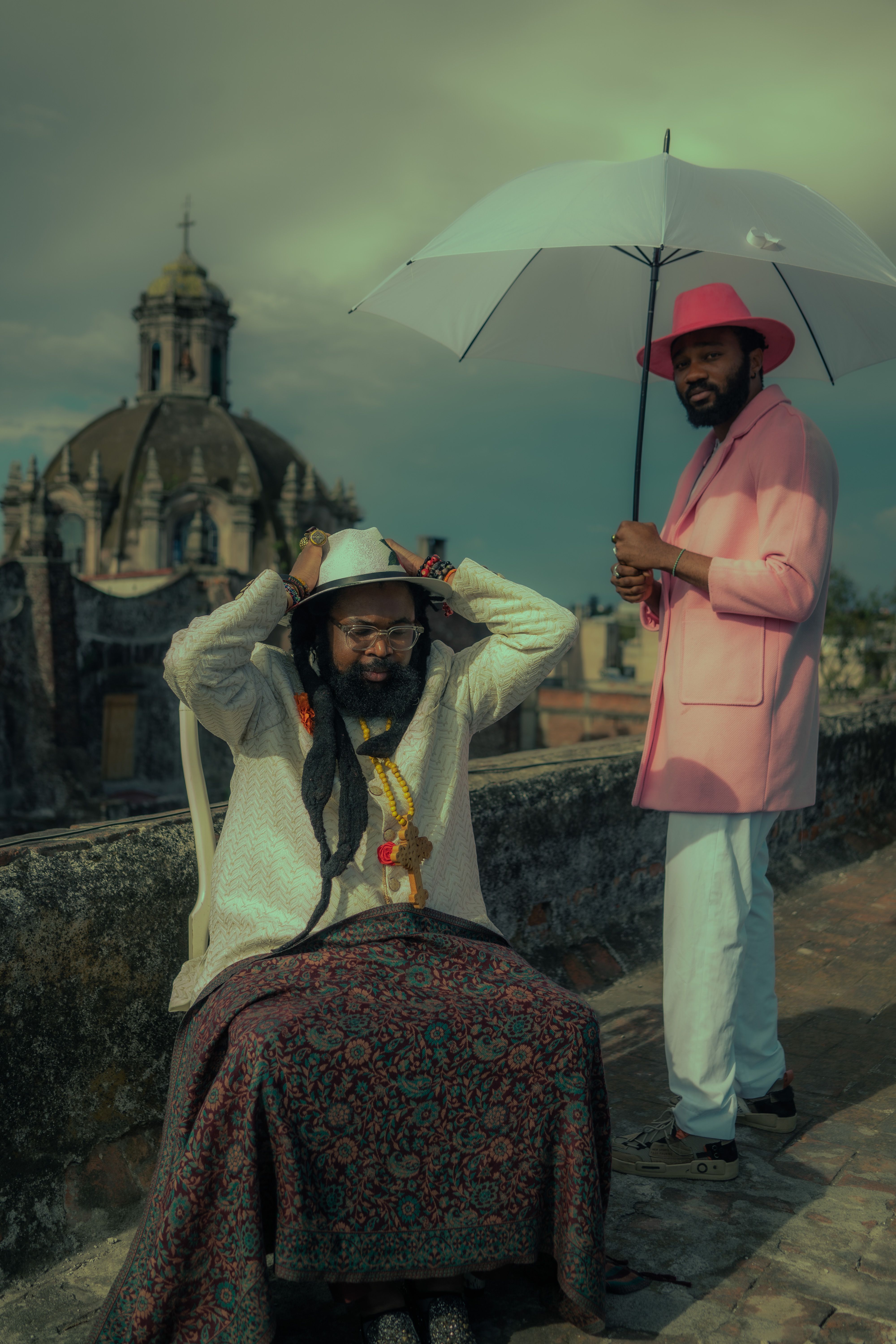 Two men in stylish attire with hats and umbrella pose on a rooftop with an antique building backdrop.