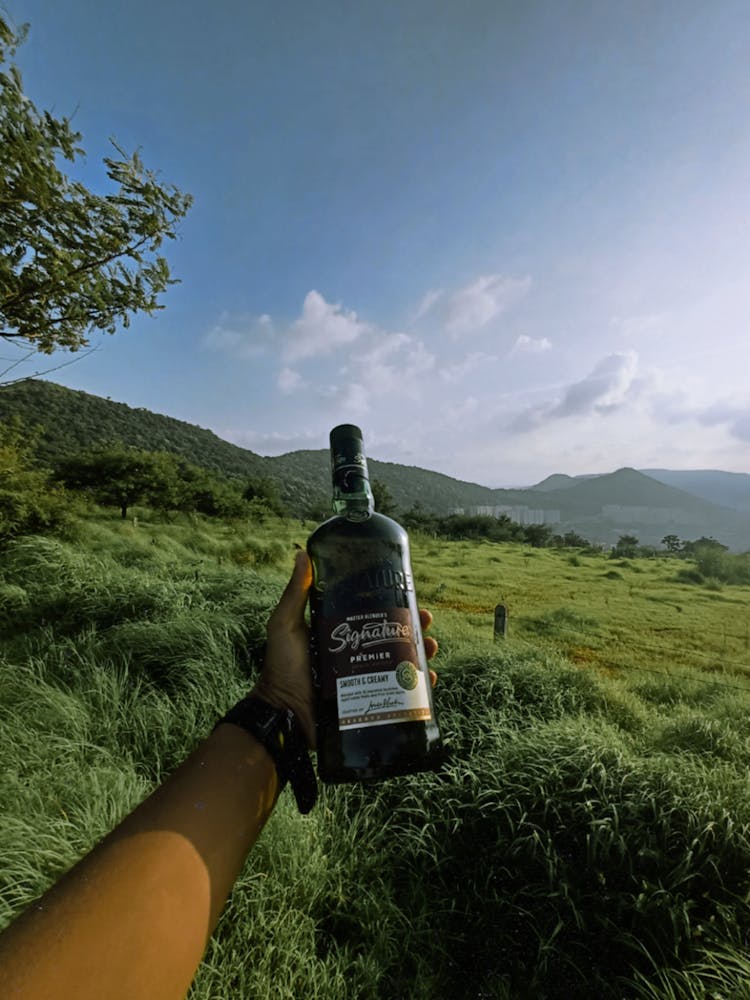 Arm Of A Person Holding A Bottle Of Whiskey Against Grassy Hills