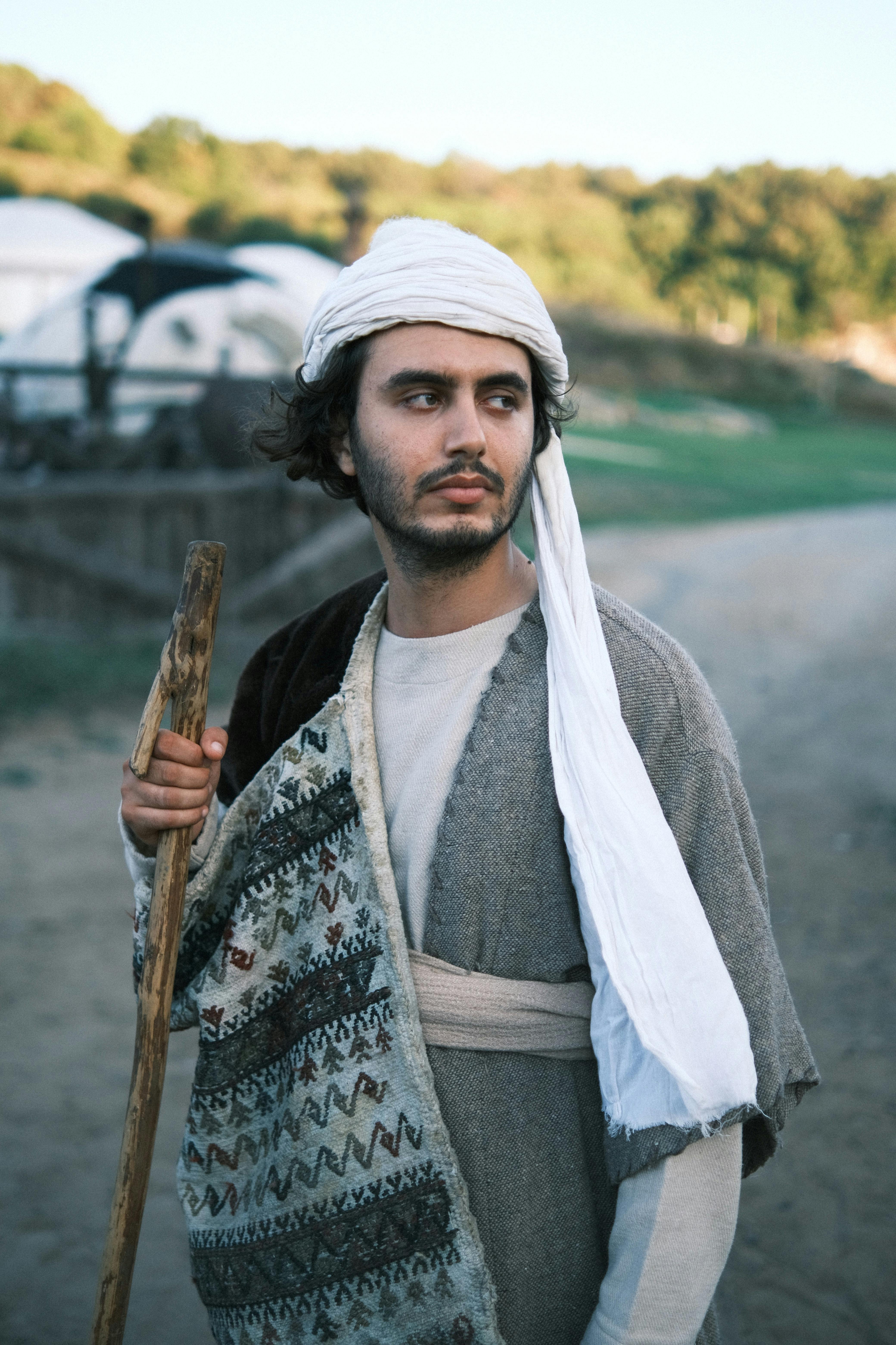 Man in traditional clothing holding a shepherd's staff in a rural setting.