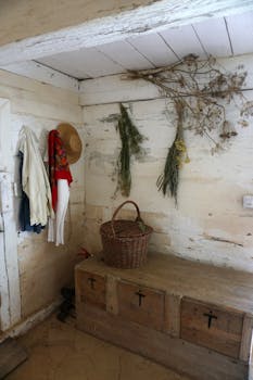 Cozy rustic interior with wooden chest, clothes, basket, and hanging herbs.