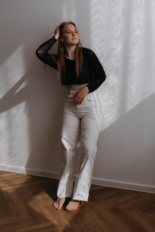 Young Blonde Woman in Black Long Sleeve Crop Top and White Pants Standing by a Wall