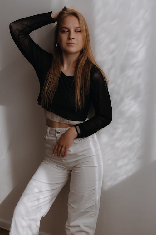Woman in White Pants and Black Shirt Posing against White Wall