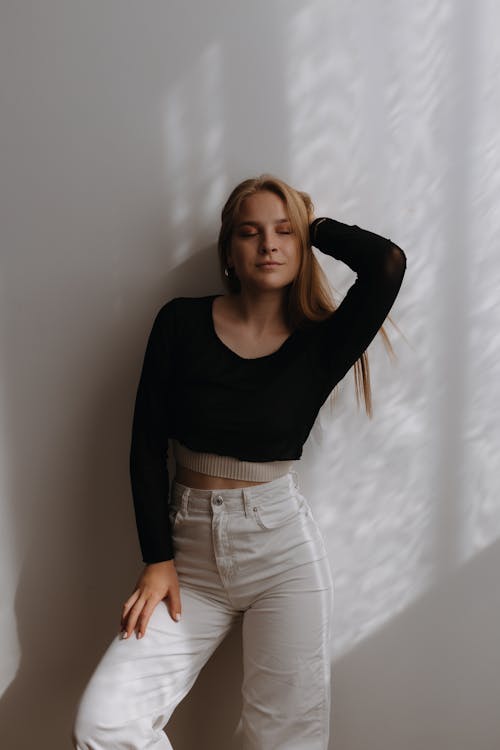 Blonde Woman in White Jeans and Black Crop Top Posing by a Grey Wall