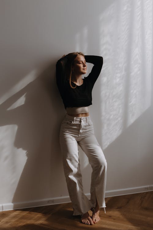 Woman in White Jeans and Black Crop Top Posing by a Wall with Hands Behind her Head