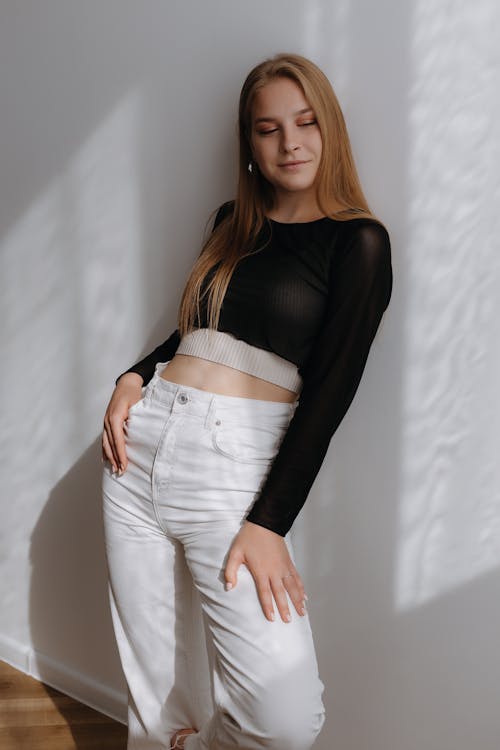 Woman in Black See Through Top and White Jeans Leaning on a Grey Wall
