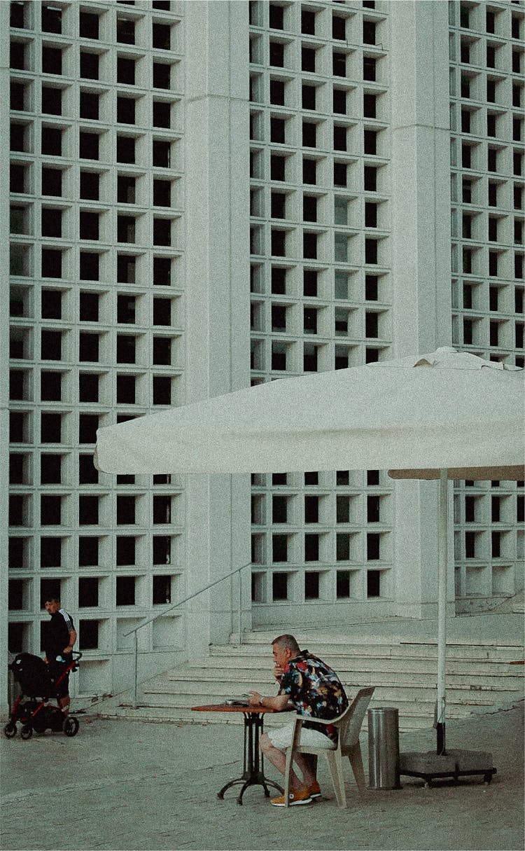 Man Sitting Under Umbrella Near White Building Wall