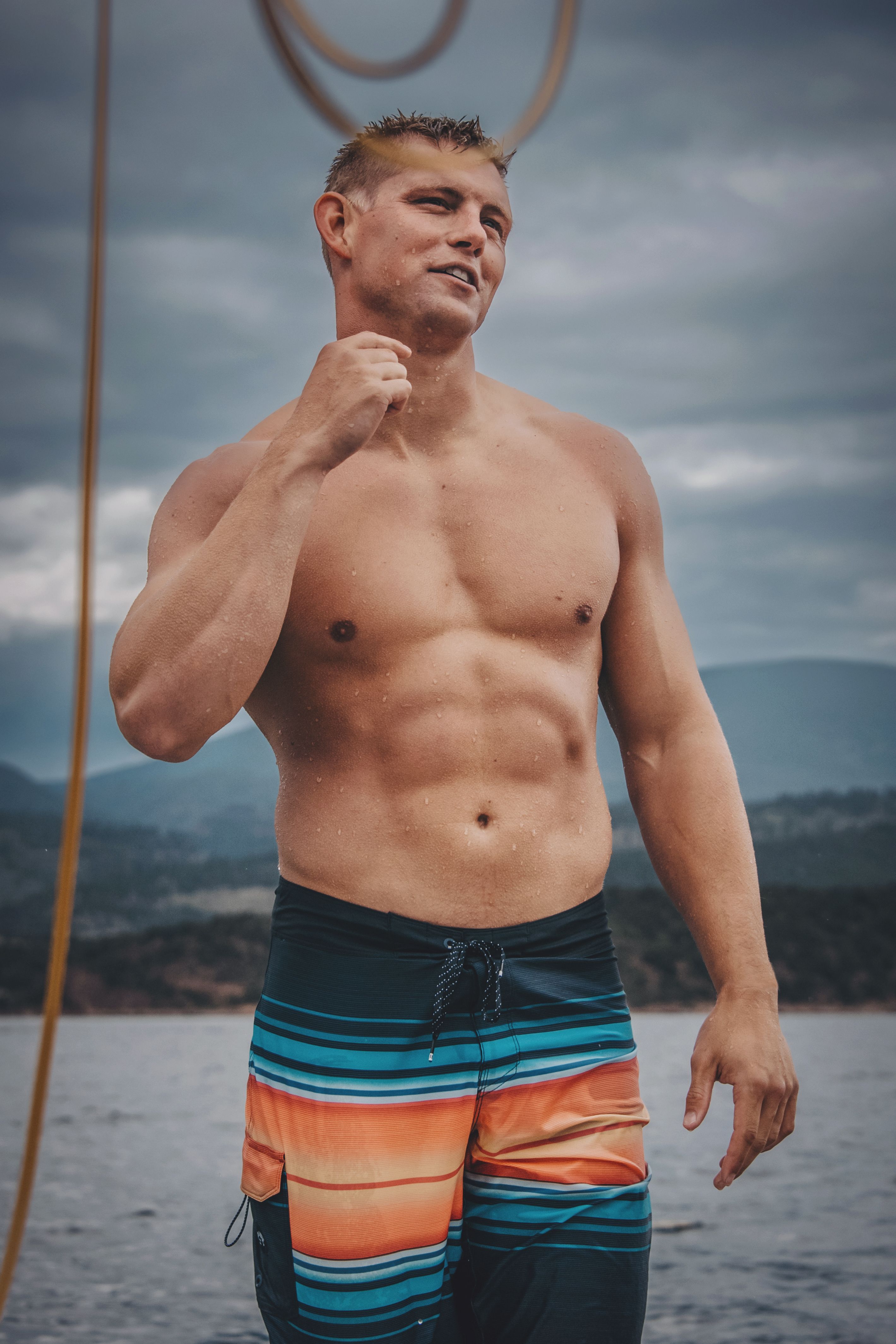 Athletic man smiling and standing confidently outdoors in swim trunks on a cloudy day.