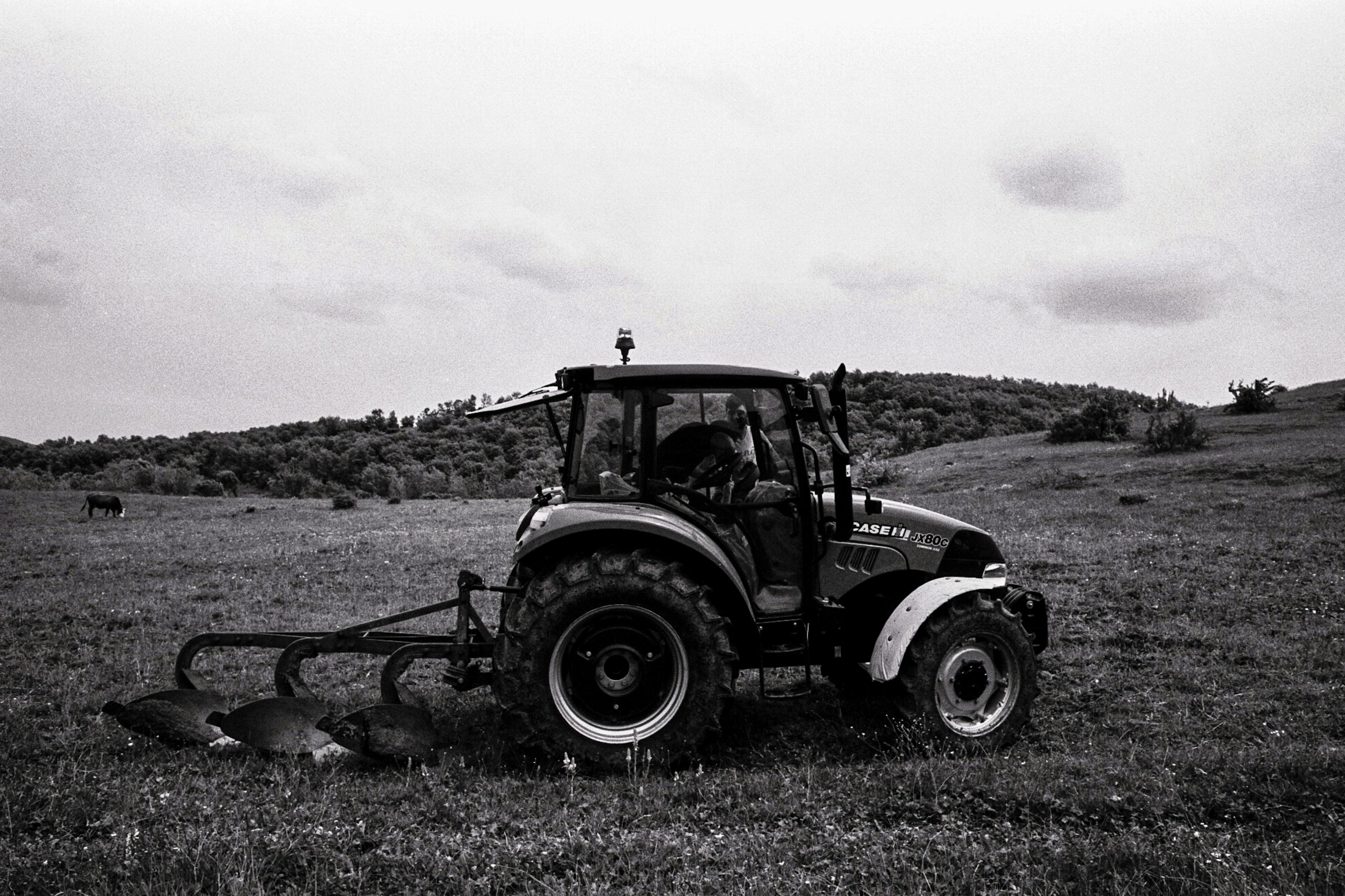 A tractor plows a field in an open countryside, set against a hilly backdrop.