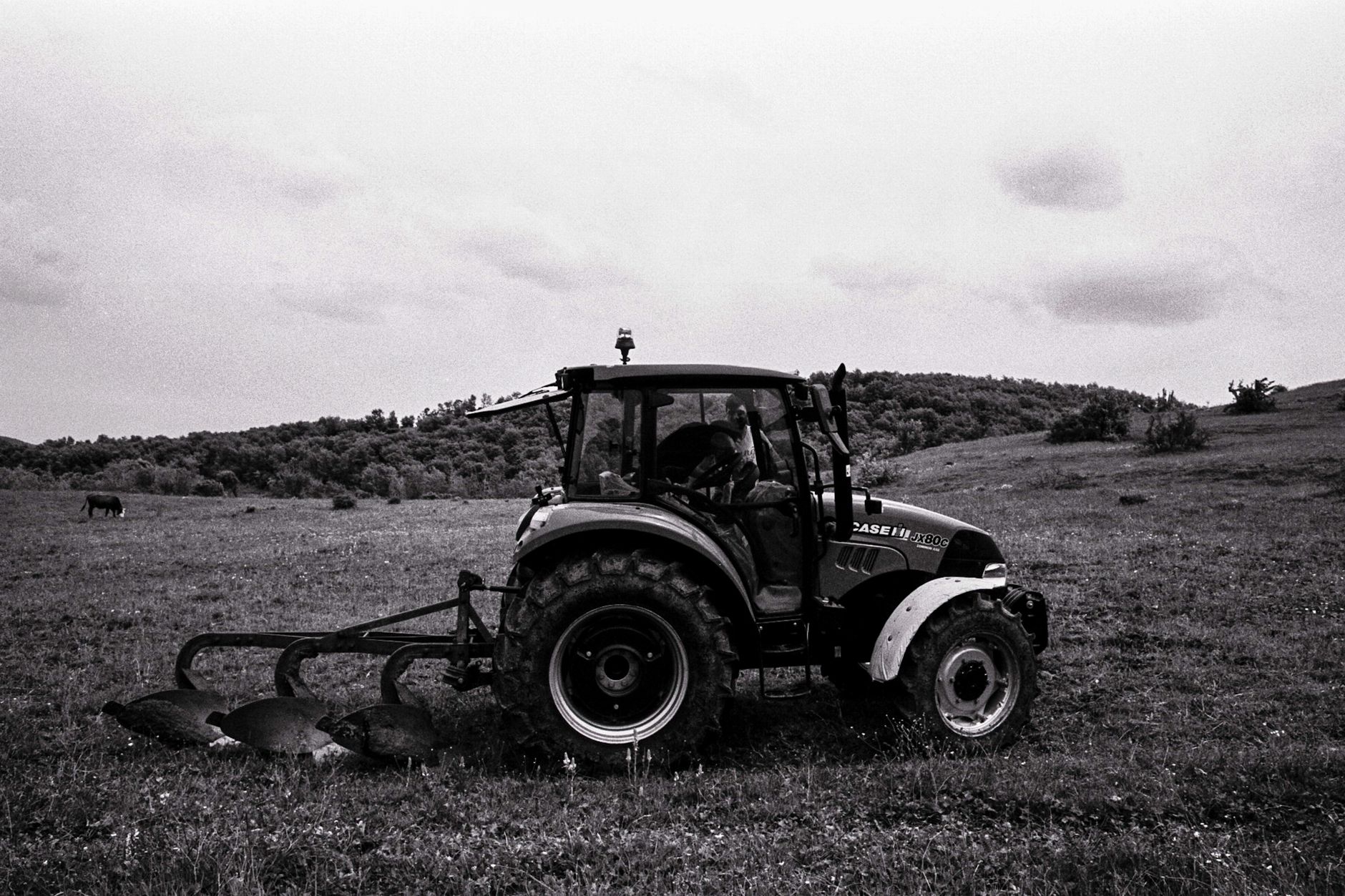 A tractor plows a field in an open countryside, set against a hilly backdrop.