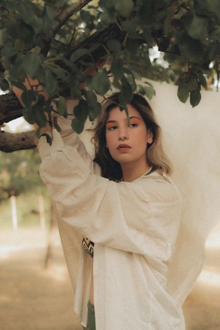Elegant portrait of a woman in a white shirt standing under a tree on a sunny day.