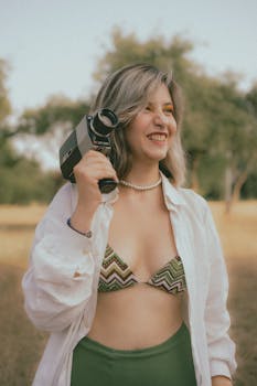 Laughing woman in bikini and shirt holding vintage camera outdoors.
