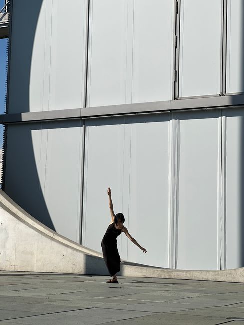 A woman dancer gracefully balancing in front of contemporary building exterior.