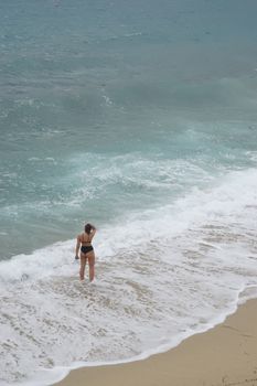 A woman in a bikini enjoying the waves on a sandy Greek beach.