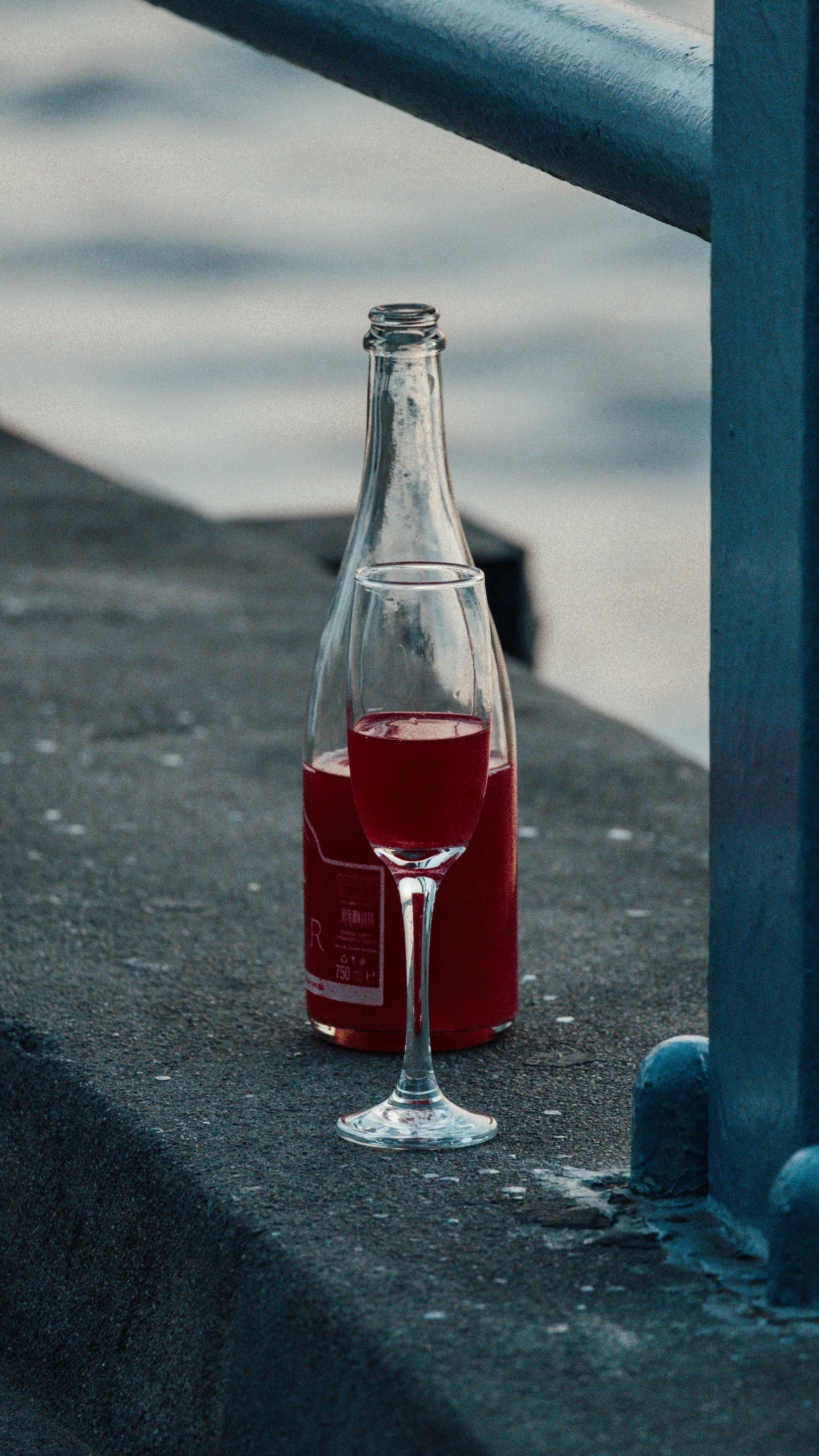 Close-up of a glass and bottle of red drink on a waterfront ledge outdoors.