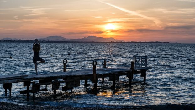 Silhouette of a woman practicing yoga on a jetty in Kos, Greece, during a picturesque sunrise.