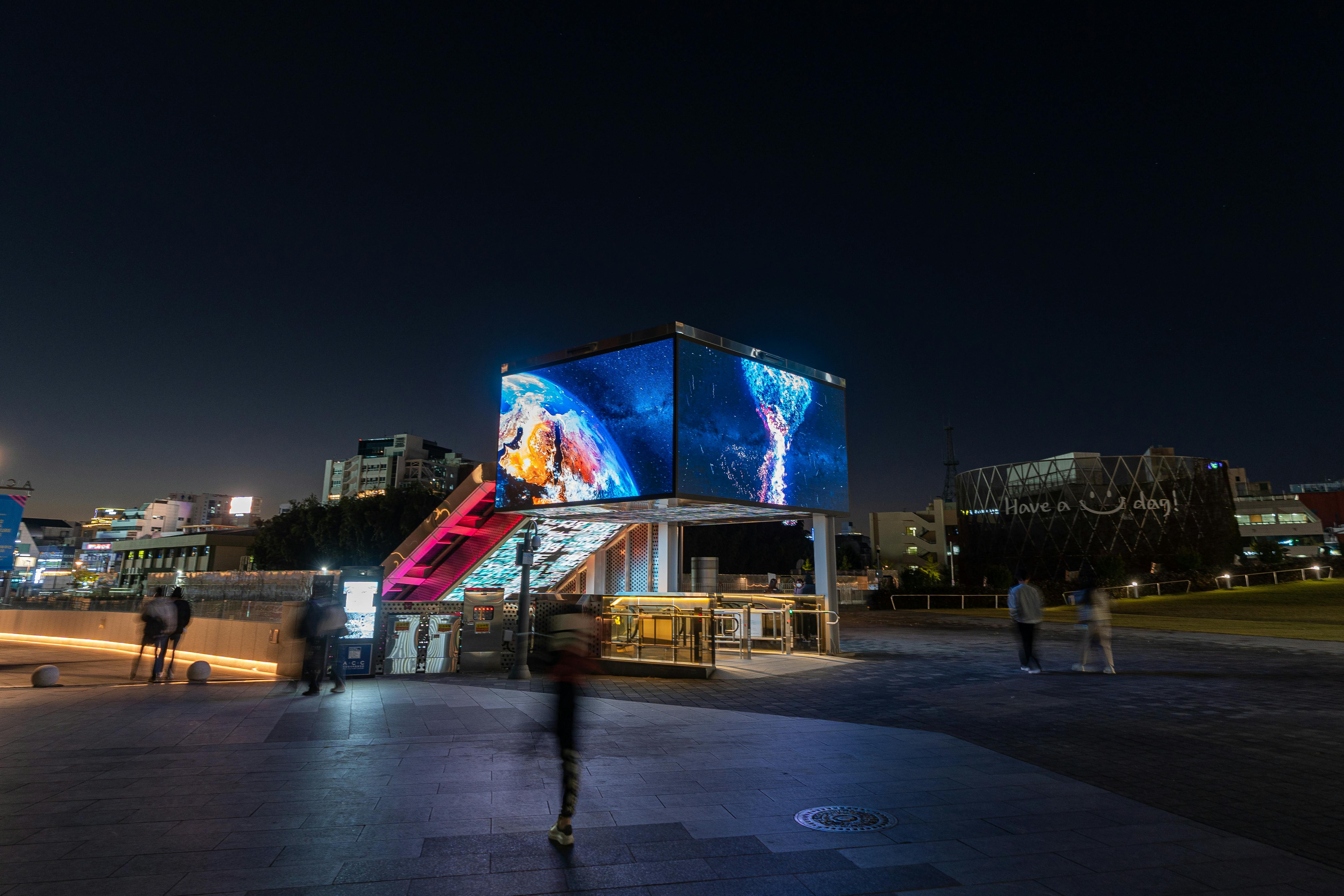 Nighttime view of a dynamic urban plaza with a vibrant digital billboard and blurred motion of people walking.